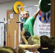 weightlifting athlete holds weights over his head as YMCA children watch