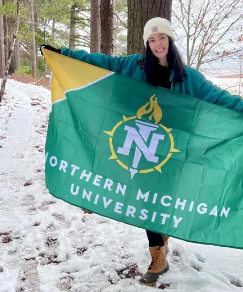 Student in a green jacket and white beanie hat standing in a snowy forest holds an NMU flag in outstretched arms