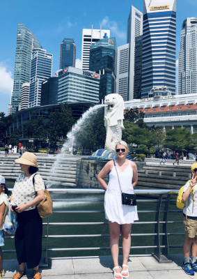 Aino Tikkenen: A person in a white dress stands in front of the Merlion fountain in Singapore, with skyscrapers in the background. It's a sunny day, and the person is smiling, wearing sunglasses, and holding a black purse.