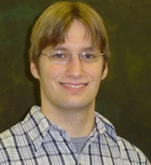 Alex Johnson-Buck: A person with glasses, light brown hair, and a friendly smile is wearing a plaid shirt. The background is a dark, muted color.