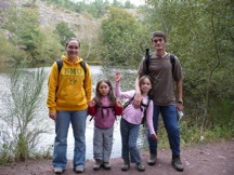 A family of four stands outdoors wearing backpacks, surrounded by trees. A body of water is visible in the background, and the sky is overcast.