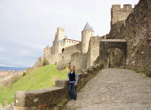 Amber Shumard: A person stands on a cobblestone path beside the imposing medieval walls and towers of Carcassonne Castle, France. The sky is overcast, and the landscape features green grass and distant hills.