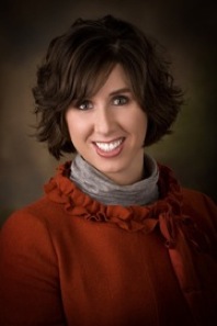 Jill Grundstrom: Woman with short brown hair smiling while wearing a red top with a ruffled neckline and a gray scarf. She is posed against a dark, neutral background.