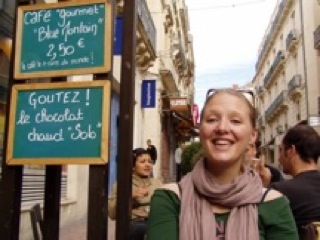 Journey Schall: A woman smiles broadly on a bustling European street. Behind her are two chalkboard signs in French advertising coffee and hot chocolate. People are walking down the narrow street lined with buildings.