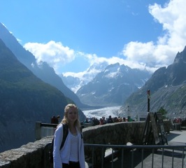 Andrea Jovanovic: A woman standing on a stone pathway overlooking a vast mountain landscape with snow-capped peaks and a cloudy sky. A crowd is visible in the background, admiring the scenery.