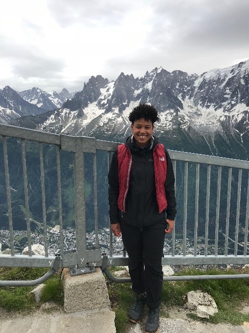 Raven Mitchell: A person stands smiling in front of a metal railing with snow-capped mountains in the background. They are wearing a red vest and dark clothing. The sky is overcast.