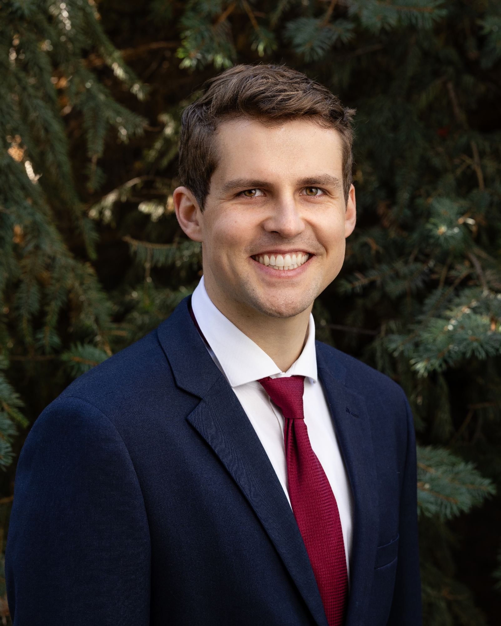 Stephen Wood: A smiling man in a dark blue suit with a white shirt and red tie stands in front of green foliage.