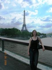 Barbara Thelen: A woman wearing a black top and jeans standing on a bridge with the Eiffel Tower visible in the background. It's a cloudy day.