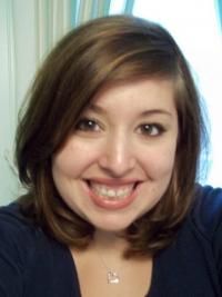 Sidney Arnold: A person with shoulder-length brown hair smiling at the camera. They are wearing a dark top and a heart-shaped necklace, with a blurred light-colored background.