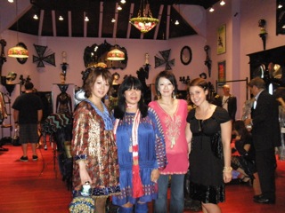 Four women pose together in a room with red flooring and eclectic decor. They wear colorful, patterned clothing, and there are various lamps and mirrors on the walls. People in the background are engaged in conversation.