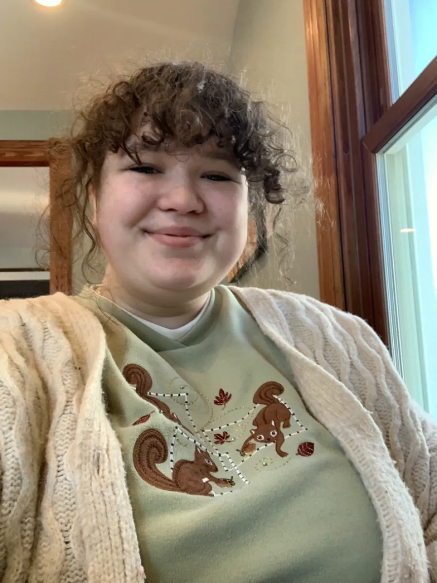 Madi smiling in a coffee shop wearing a green sweatshirt that has squirrels frolicking with acorns on it